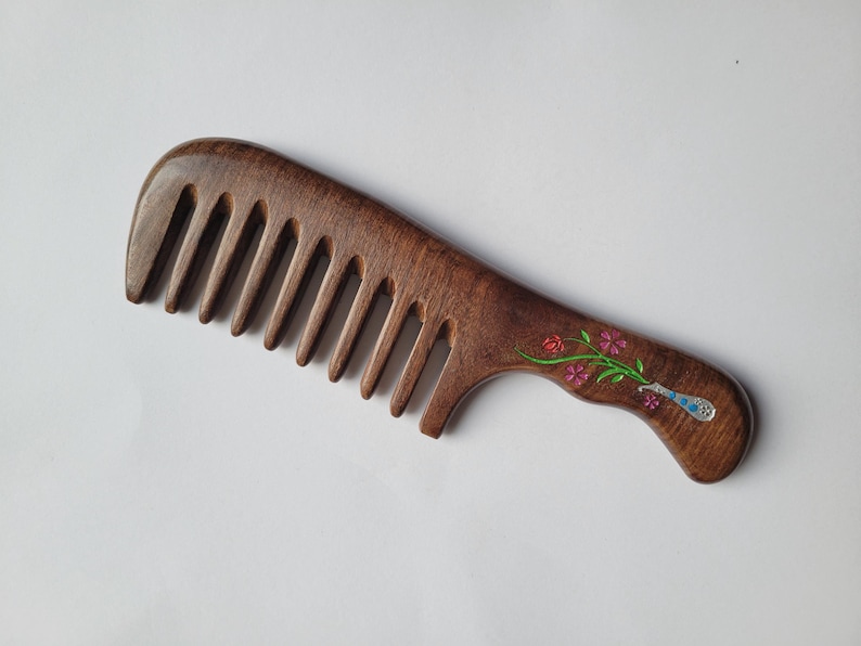 May include: A dark brown wooden wide-tooth comb featuring a smooth, curved handle. The handle is decorated with an inlaid design of a silver-toned vase containing a small green stem with pink and red flowers and blue accents.