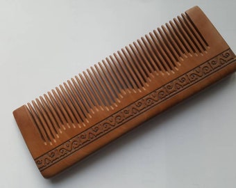 Pode incluir: Um pente de madeira castanho com um padrão decorativo esculpido no cabo. O pente tem dentes finos e é projetado para desembaraçar o cabelo.