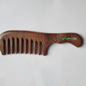 May include: A dark brown wooden hair comb with wide teeth and a curved handle. The handle features an inlaid decorative design of a green stem with small pink flowers and a blue and silver patterned accent near the end of the handle.