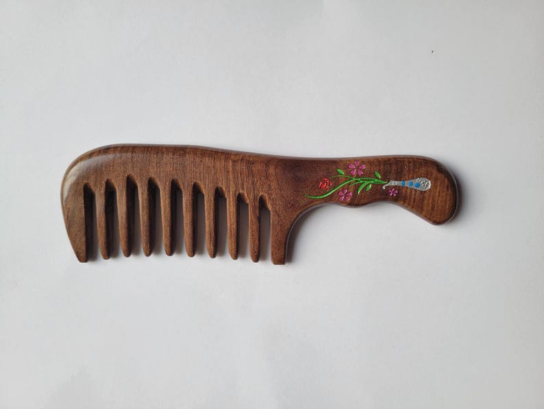 May include: A dark brown wooden hair comb with wide teeth and a curved handle. The handle features an inlaid decorative design of a green stem with small pink flowers and a blue and silver patterned accent near the end of the handle.