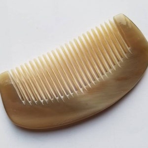 Könnte beinhalten: Ein hellbrauner Kamm mit eng stehenden Zähnen. Der Kamm hat eine gebogene Form und besteht aus einem Material, das wie Horn oder eine ähnliche natürliche Substanz aussieht. Der Kamm ist vor einem weißen Hintergrund platziert.