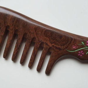 A dark brown wooden comb with a floral design on the handle. The flowers are pink and green, and the handle has a silver-colored accent.