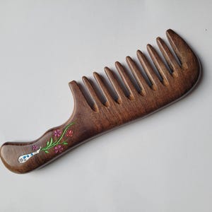 May include: A dark brown wooden wide-tooth comb featuring a smooth, polished finish. The handle is decorated with a small, painted floral design consisting of green stems, pink flowers, and a white and blue patterned bulb-shaped element.