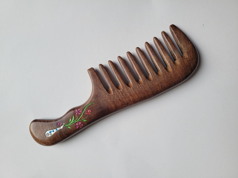May include: A dark brown wooden wide-tooth comb featuring a smooth, polished finish. The handle is decorated with a small, painted floral design consisting of green stems, pink flowers, and a white and blue patterned bulb-shaped element.