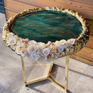 Könnte beinhalten: Ein runder Beistelltisch mit einem goldfarbenen Metallgestell und einer einzigartigen Tischplatte. Die Tischplatte hat ein tiefgrünes, marmoriertes Design mit einem goldfarbenen, strukturierten Rand. Der Rand ist mit weißen und bernsteinfarbenen Kristallen verziert.