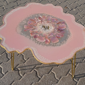 Peut inclure: Une table basse en résine rose avec une forme organique unique et un centre transparent rempli de cristaux roses et blancs. La table est soutenue par deux pieds en épingle à cheveux dorés.