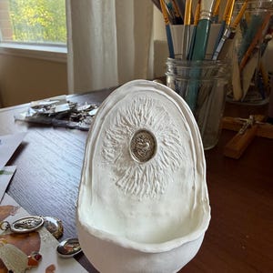 May include: A white, sculpted religious art piece with a textured surface and a central emblem. The piece is set on a wooden surface, with paintbrushes and other art supplies visible in the background.