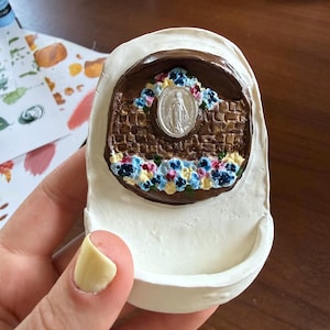 May include: A white, sculpted religious item with a brown, brick-like archway. The archway features a silver medallion and is adorned with blue, yellow, and pink flowers. The item is held in a hand.