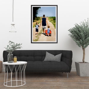 K&ouml;nnte beinhalten: Gerahmtes Foto einer Familie, die einen Feldweg entlanggeht. Ein Mann in einem &auml;rmellosen Hemd und Shorts h&auml;lt die H&auml;nde von zwei Kindern. Ein Kind sitzt in einem roten Spielzeugwagen. Das Foto wird an einer wei&szlig;en Wand gezeigt.