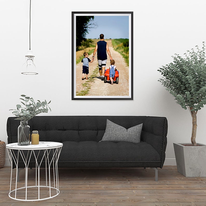 K&ouml;nnte beinhalten: Gerahmtes Foto einer Familie, die einen Feldweg entlanggeht. Ein Mann in einem &auml;rmellosen Hemd und Shorts h&auml;lt die H&auml;nde von zwei Kindern. Ein Kind sitzt in einem roten Spielzeugwagen. Das Foto wird an einer wei&szlig;en Wand gezeigt.