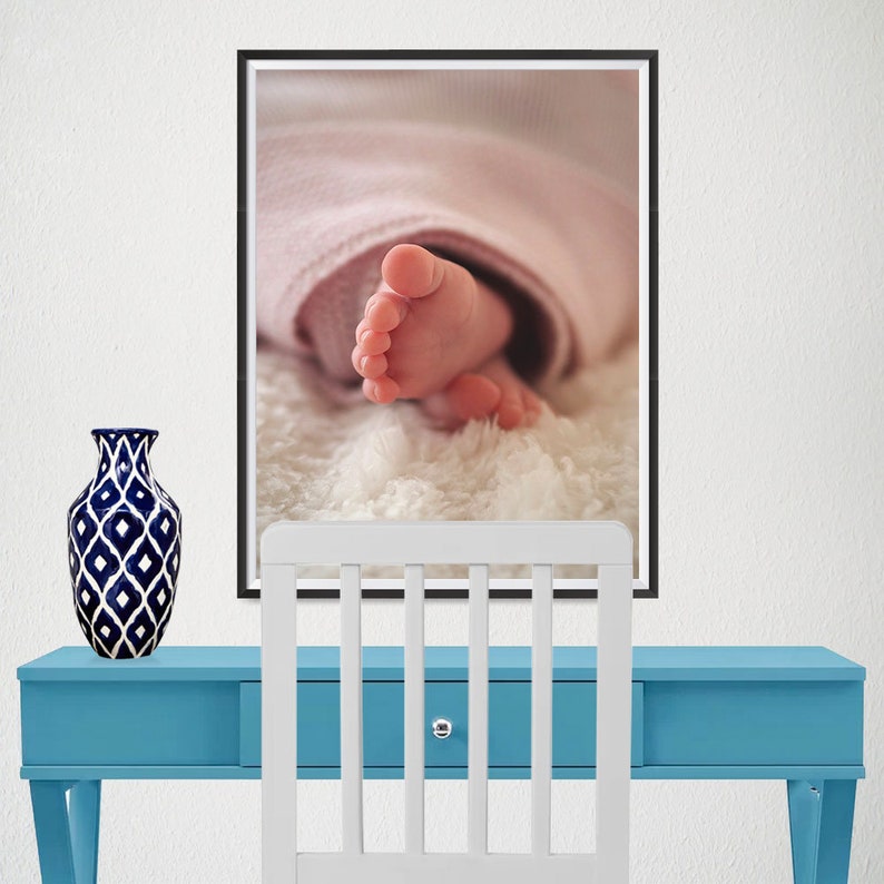 K&ouml;nnte beinhalten: Gerahmtes Foto von Babyf&uuml;&szlig;en, die Zehen sind sichtbar. Die F&uuml;&szlig;e sind rosa und liegen auf einer flauschigen wei&szlig;en Oberfl&auml;che, eingeh&uuml;llt in eine rosa Decke. Das Foto h&auml;ngt &uuml;ber einem blauen Schreibtisch mit einem wei&szlig;en Stuhl und einer dekorativen Vase.