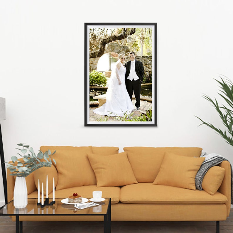K&ouml;nnte beinhalten: Ein gerahmtes Hochzeitsfoto eines Brautpaares. Die Braut tr&auml;gt ein wei&szlig;es Brautkleid und einen Schleier, der Br&auml;utigam einen schwarzen Smoking. Das Foto h&auml;ngt &uuml;ber einem gelben Sofa mit Kissen, einem Glastisch und einer Vase mit Blumen.
