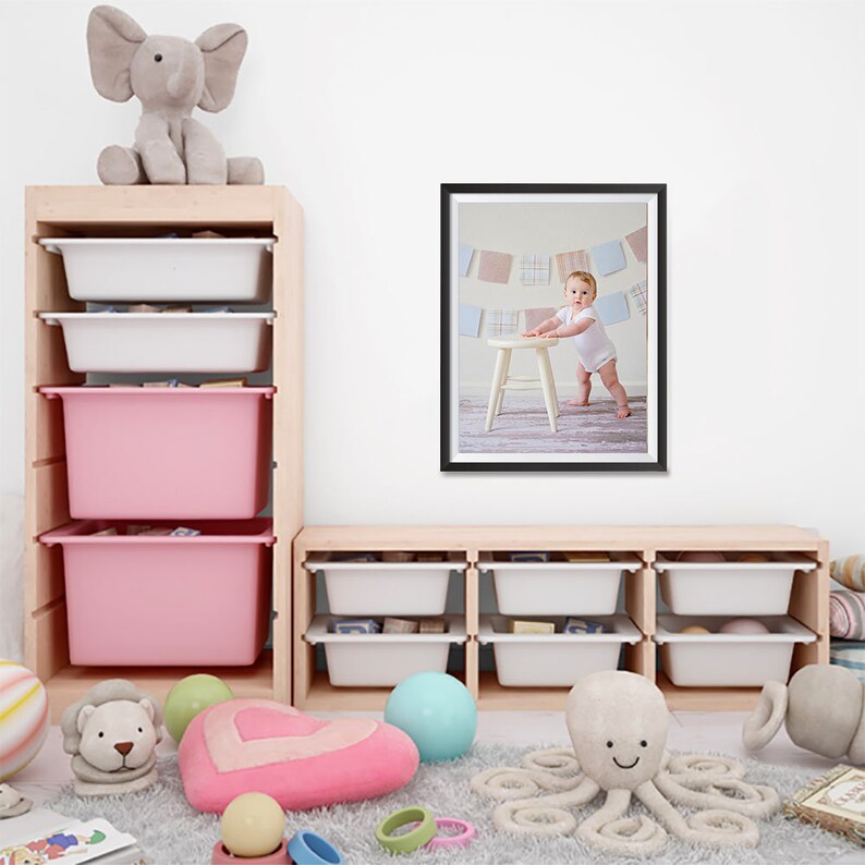 K&ouml;nnte beinhalten: Holzaufbewahrungseinheiten mit wei&szlig;en und rosa Beh&auml;ltern, ein gerahmtes Foto eines Babys und Pl&uuml;schtiere. Zu den Spielzeugen geh&ouml;ren ein grauer Elefant, ein L&ouml;we, ein herzf&ouml;rmiges Kissen, ein blauer Ball und ein Oktopus.