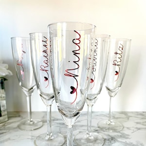 May include: Set of five clear glass champagne flutes with personalised names in pink script. The names are "Rita", "Emma", "Mina", "Rachael", and "Lily".
