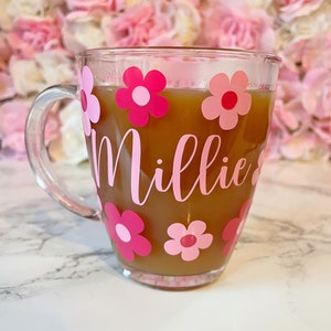 May include: A clear glass mug with a pink handle and pink flowers and the name "Millie" written on it. The mug is filled with a brown liquid.