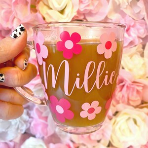 May include: A clear glass mug with a pink and white floral design and the name "Millie" written in pink. The mug is filled with a brown liquid and is being held by a hand.