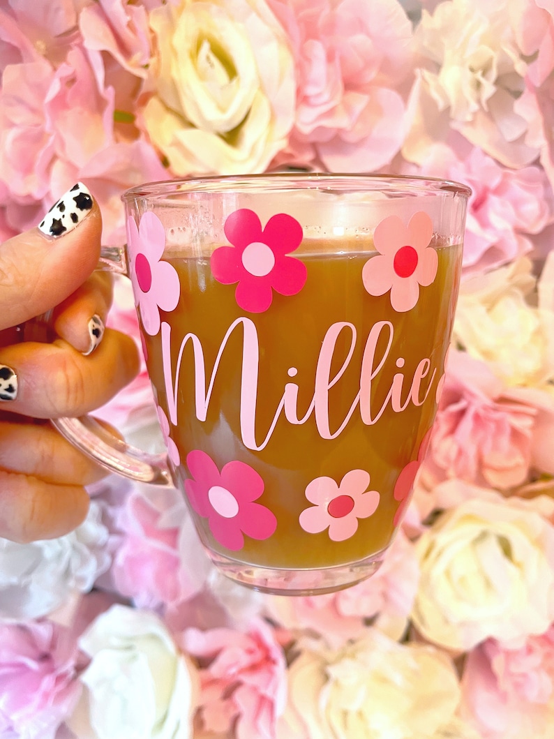 May include: A clear glass mug with a pink and white floral design and the name "Millie" written in pink. The mug is filled with a brown liquid and is being held by a hand.