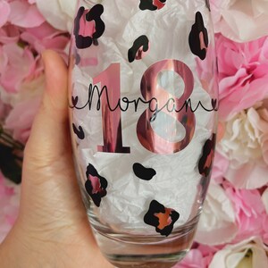May include: A clear glass with a pink and black leopard print design. The glass has the number 18 in rose gold foil and the name "Morgan" written in black cursive.