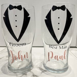 May include: Two clear glass pint glasses with black tuxedo designs and the words "Groom John" and "Best Man Paul" in rose gold lettering.