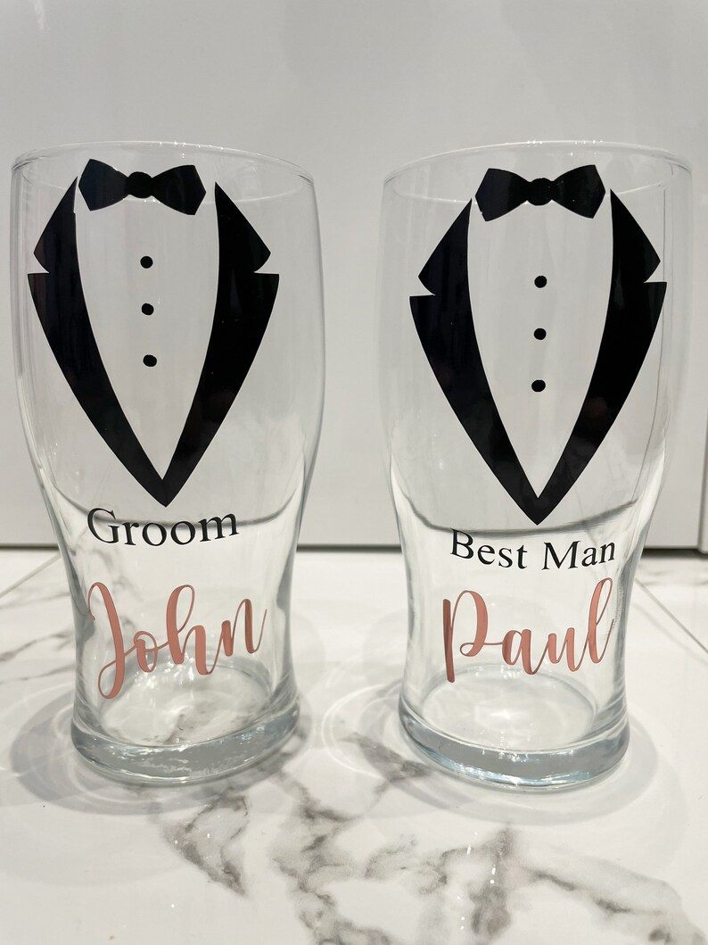 May include: Two clear glass pint glasses with black tuxedo designs and the words "Groom John" and "Best Man Paul" in rose gold lettering.