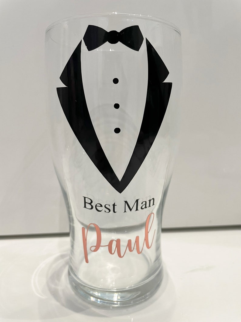 May include: A clear glass pint glass with a black tuxedo design and the text "Best Man Paul" in rose gold lettering.