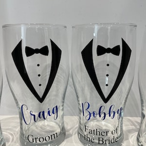 May include: Two clear glass pint glasses with black tuxedo designs and blue text. The glass on the left says "Craig Groom" and the glass on the right says "Bobby Father of the Bride".