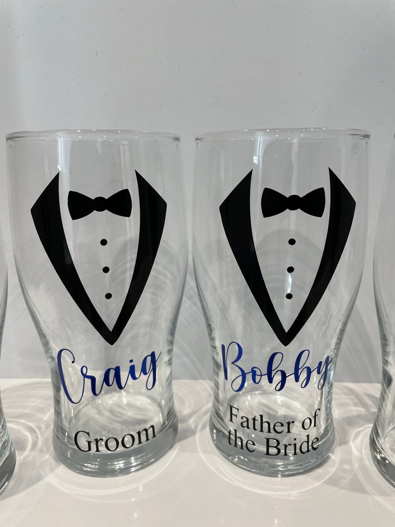 May include: Two clear glass pint glasses with black tuxedo designs and blue text. The glass on the left says "Craig Groom" and the glass on the right says "Bobby Father of the Bride".