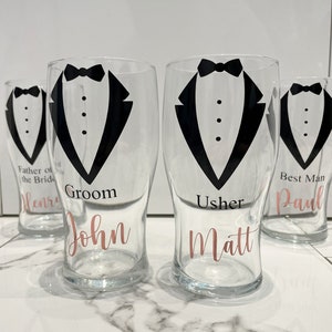 May include: Four clear glass pint glasses with black tuxedo designs and rose gold lettering. The glasses are labelled "Groom John", "Usher Matt", "Best Man Paul", and "Father of the Bride Henry".