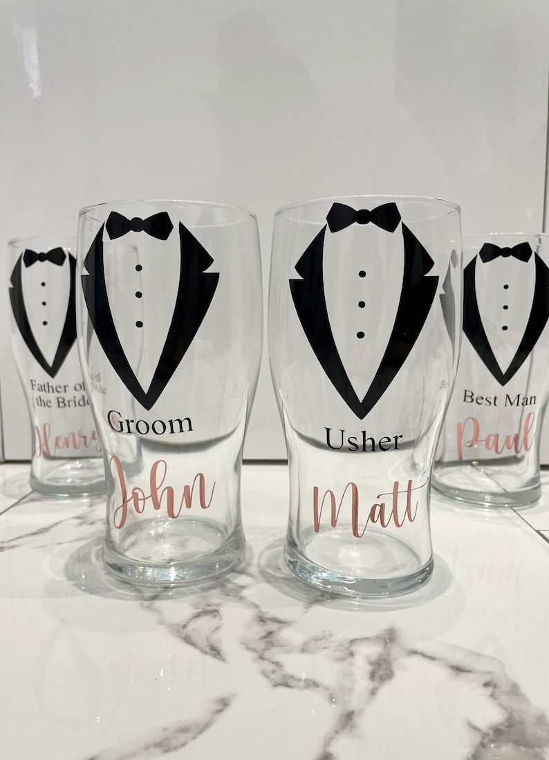 May include: Four clear glass pint glasses with black tuxedo designs and rose gold lettering. The glasses are labelled "Groom John", "Usher Matt", "Best Man Paul", and "Father of the Bride Henry".