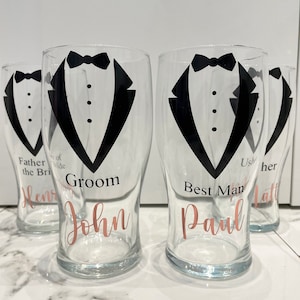 May include: Four clear glass pint glasses with black tuxedo designs and rose gold lettering. The glasses are labelled "Groom John", "Best Man Paul", "Father of the Bride Henry", and "Usher Matt".