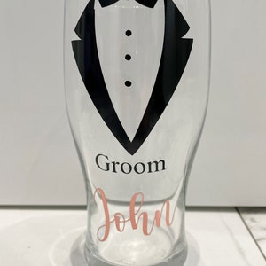 May include: A clear glass with a black tuxedo design and the words "Groom John" in pink lettering.