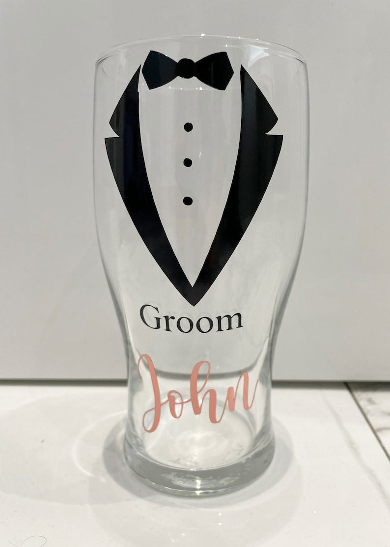 May include: A clear glass with a black tuxedo design and the words "Groom John" in pink lettering.