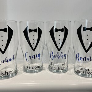 May include: Four clear glass pint glasses with black tuxedo designs and blue text. The glasses are labelled "Michael Usher", "Craig Groom", "Bobby Father of the Bride", and "Ronnie Father of the Groom".