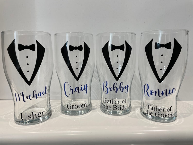 May include: Four clear glass pint glasses with black tuxedo designs and blue text. The glasses are labelled "Michael Usher", "Craig Groom", "Bobby Father of the Bride", and "Ronnie Father of the Groom".