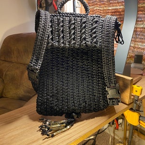 May include: A black paracord backpack with a woven design and a black strap. The backpack has a small tassel with gold beads that spell out "PLEASE" and "BE KIND".