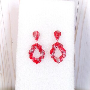 May include: A pair of red and silver dangle earrings. Each earring features a teardrop-shaped top and a larger, open teardrop design below. The earrings have a marbled appearance with silver accents.
