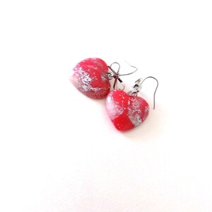May include: A pair of heart-shaped earrings. Each earring features a red base with silver-colored accents. The earrings have a hook closure and are set against a white background.