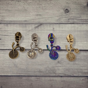 May include: Four different colored metal zipper pulls shaped like a vintage tricycle. The zipper pulls are in gold, silver, rainbow, and bronze.
