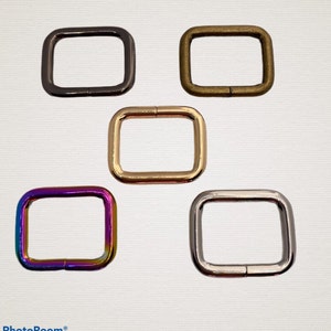 May include: Five metal square rings in different colors: black, gold, silver, brass, and iridescent. The rings are all the same size and shape.