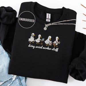 Könnte beinhalten: Schwarzes Sweatshirt mit Rundhalsausschnitt und gesticktem Enten-Design. Der Text "doing social worker stuff" ist darunter gedruckt. Eine silberne Halskette und zerrissene Jeans sind ebenfalls im Bild zu sehen.