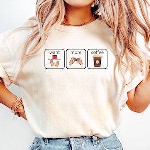 May include: A cream-coloured short-sleeve t-shirt featuring three rectangular boxes with black outlines. Each box contains a graphic illustration and text: 'want' with hands, 'more' with hands, and 'coffee' with a brown coffee cup icon.