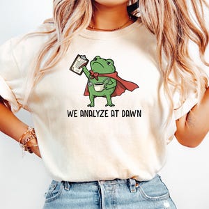 Peut inclure: Un t-shirt couleur crème avec une illustration d'une grenouille verte portant une cape rouge et un stéthoscope, tenant un presse-papier et une tasse de café. Sous la grenouille, le texte WE ANALYZE AT DAWN est écrit en lettres capitales noires.
