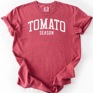 May include: A crimson short-sleeved t-shirt featuring the white text TOMATO SEASON printed in a bold, arched collegiate font across the chest. The garment has a crew neckline and a standard fit, with the word Crimson written in the bottom corner.