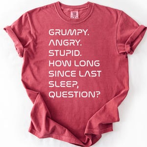 May include: Crimson-colored t-shirt with the text "GRUMPY. ANGRY. STUPID. HOW LONG SINCE LAST SLEEP, QUESTION?" in white block letters. The shirt has rolled-up sleeves and a crew neck.