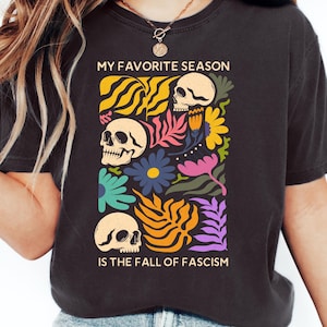 以下が含まれることがあります： 鮮やかなグラフィックが施されたダークグレーのTシャツ。グラフィックには3つのスカル、花、そして「MY FAVORITE SEASON IS THE FALL OF FASCISM」というテキストが描かれています。デザインには、黄色、オレンジ、青、紫の要素が含まれています。