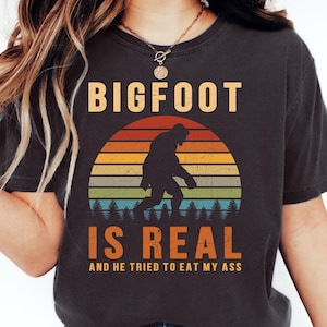Op de afbeelding: Donkergrijs t-shirt met de tekst "BIGFOOT IS REAL" in oranje en wit. Het ontwerp toont een Bigfoot-silhouet voor een zonsondergang met oranje, gele, groene en blauwe strepen. De tekst "AND HE TRIED TO EAT MY ASS" staat eronder.