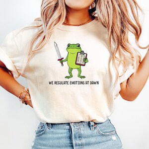 Op de afbeelding: Een crèmekleurig T-shirt met korte mouwen en een afbeelding van een groene kikker die een zwaard en een klembord vasthoudt. De tekst onder de kikker luidt "WE REGULATE EMOTIONS AT DAWN". Het T-shirt wordt gedragen met een lichtblauwe spijkerbroek.