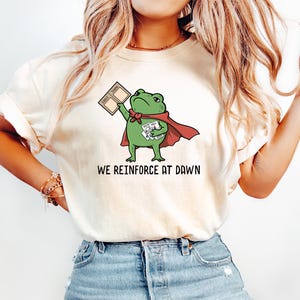 May include: A cream-colored short-sleeve t-shirt featuring a graphic of a green frog wearing a red cape. The frog holds a 'FIRST THEN' board and a stack of cards. Below the illustration, the text reads 'WE REINFORCE AT DAWN' in black letters.