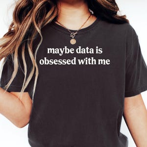 Pode incluir: T-shirt cinza escuro com o texto branco "maybe data is obsessed with me". A camisa é de gola redonda e parece ser feita de um material macio. A modelo está usando jeans azul e um colar de ouro.