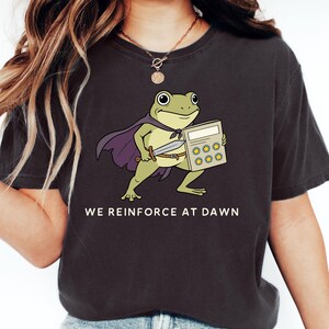 Könnte beinhalten: Dunkelgraues T-Shirt mit einer Cartoon-Froschfigur, die einen Umhang trägt und einen Taschenrechner und ein Schwert hält. Der Text "WE REINFORCE AT DAWN" ist unter der Frosch-Illustration aufgedruckt. Das T-Shirt hat einen bequemen, lässigen Stil.